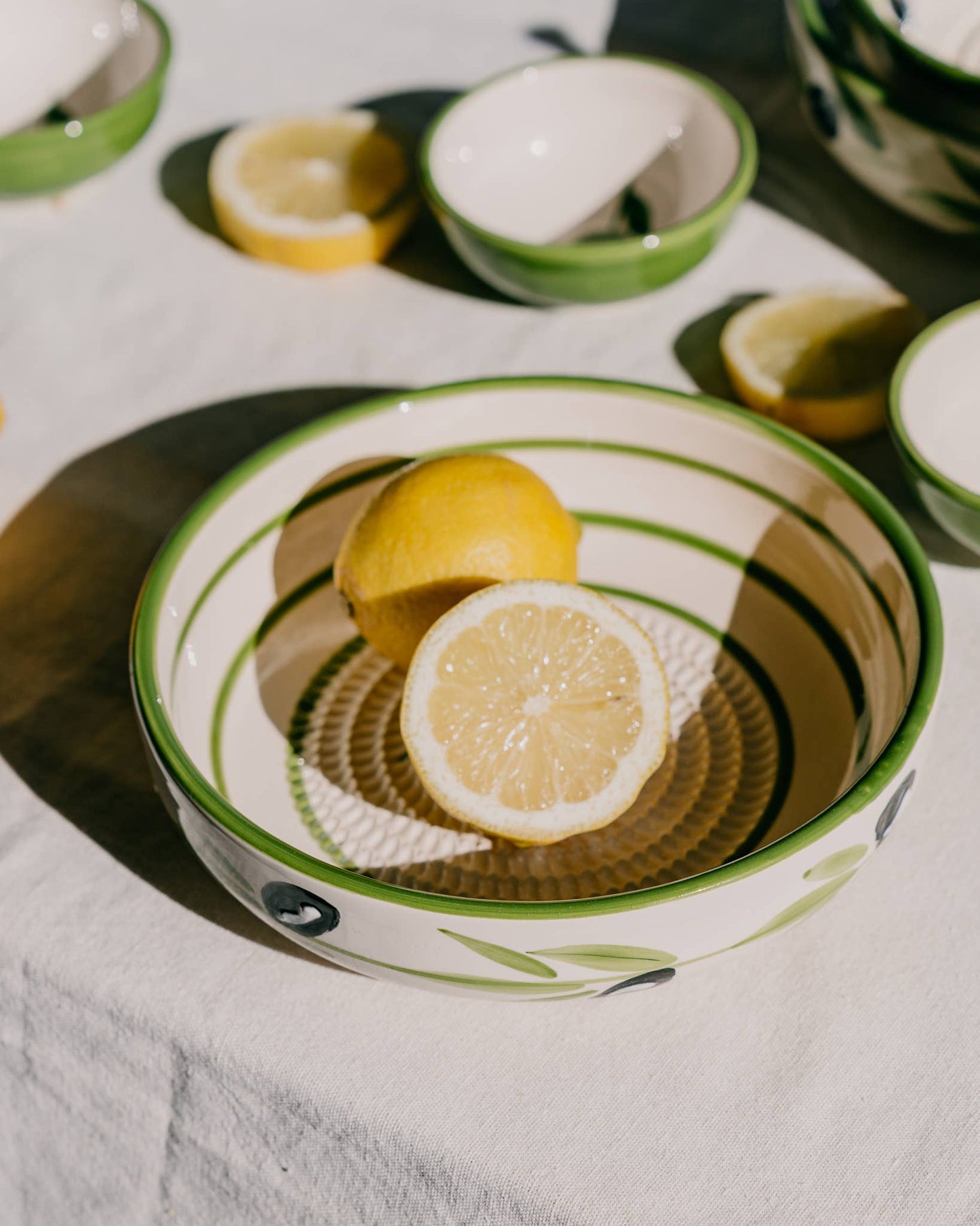 Die Alfar Tierra Cocida | Große Keramik-Reibplatte „Olive“ von Alfar Tierra Cocida dient als farbenfroher Blickfang: Die handbemalte Keramikplatte, verziert mit grünen und schwarzen Olivenmotiven, bietet Platz für eine ganze Zitrone und eine halbe Zitrone. Weitere Schälchen und frische Zitronenscheiben sind auf einer sonnendurchfluteten weißen Tischdecke verteilt und fangen den charmanten Geist der mediterranen Küchenhilfe ein.