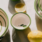 Handgefertigt in Spanien: Die kleine Keramikschale „Olive“ von Alfar Tierra Cocida mit grünem Rand und filigranen Blattmotiven ruht auf einem hellen Tuch, neben einer Zitrone und einer Zitronenhälfte. Das mediterrane Design der Keramikschale von Alfar Tierra Cocida verleiht der Szene durch das sanfte Spiel von Licht und Schatten sommerliche Leichtigkeit.