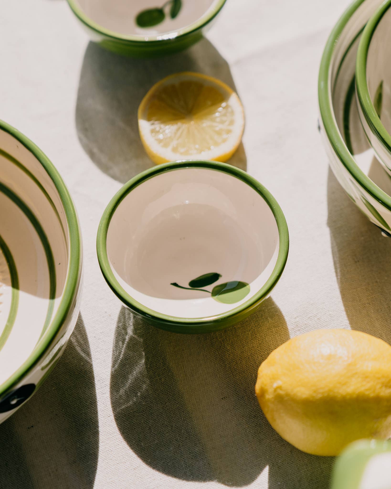 Handgefertigt in Spanien: Die kleine Keramikschale „Olive“ von Alfar Tierra Cocida mit grünem Rand und filigranen Blattmotiven ruht auf einem hellen Tuch, neben einer Zitrone und einer Zitronenhälfte. Das mediterrane Design der Keramikschale von Alfar Tierra Cocida verleiht der Szene durch das sanfte Spiel von Licht und Schatten sommerliche Leichtigkeit.
