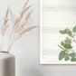 Der Wildblumen Atelier | Wandkalender 2026 mit handgezeichneten Blumen, FSC, hängt neben einer weißen Vase mit getrockneten Grashalmen vor einer hellen Wand.