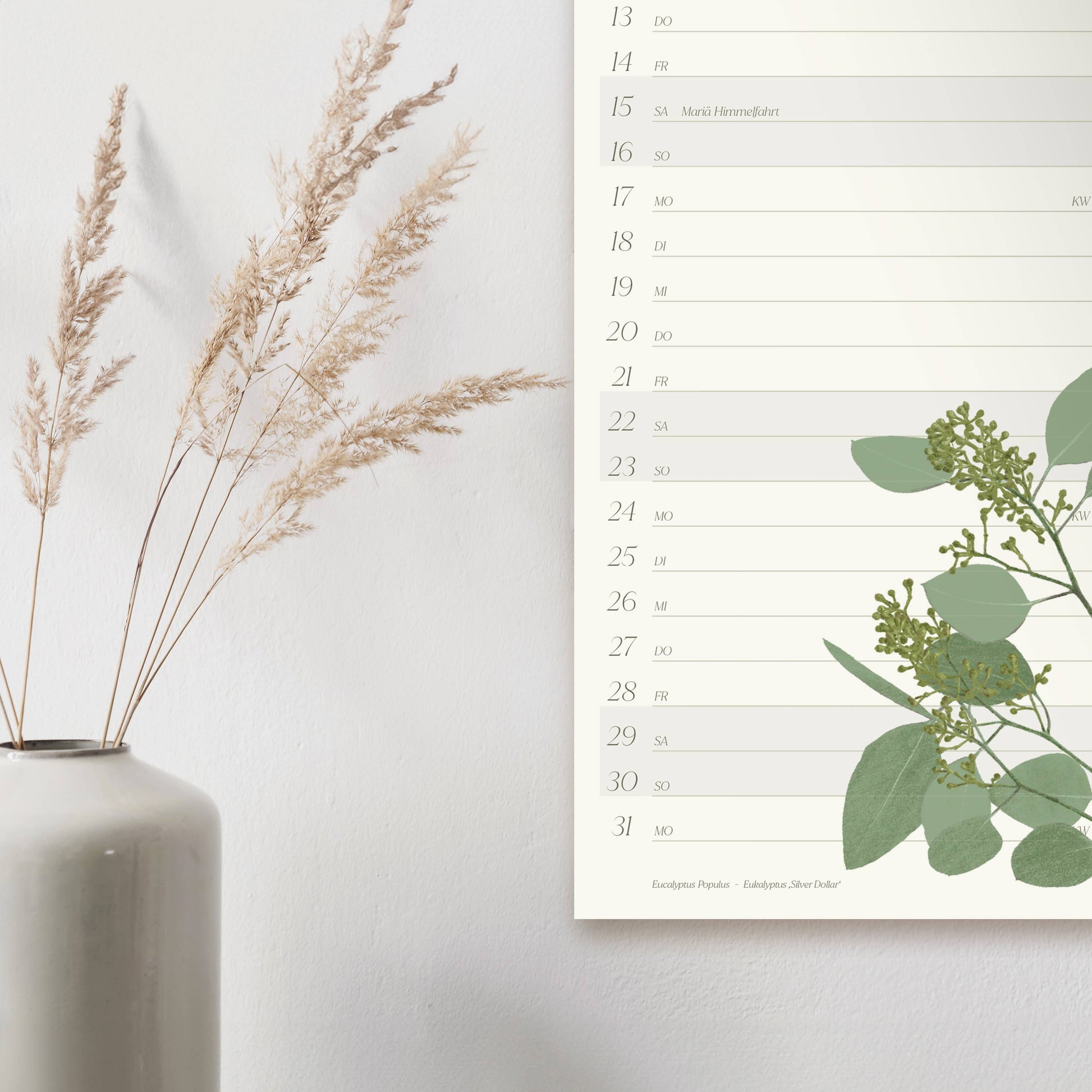 Der Wildblumen Atelier | Wandkalender 2026 mit handgezeichneten Blumen, FSC, hängt neben einer weißen Vase mit getrockneten Grashalmen vor einer hellen Wand.