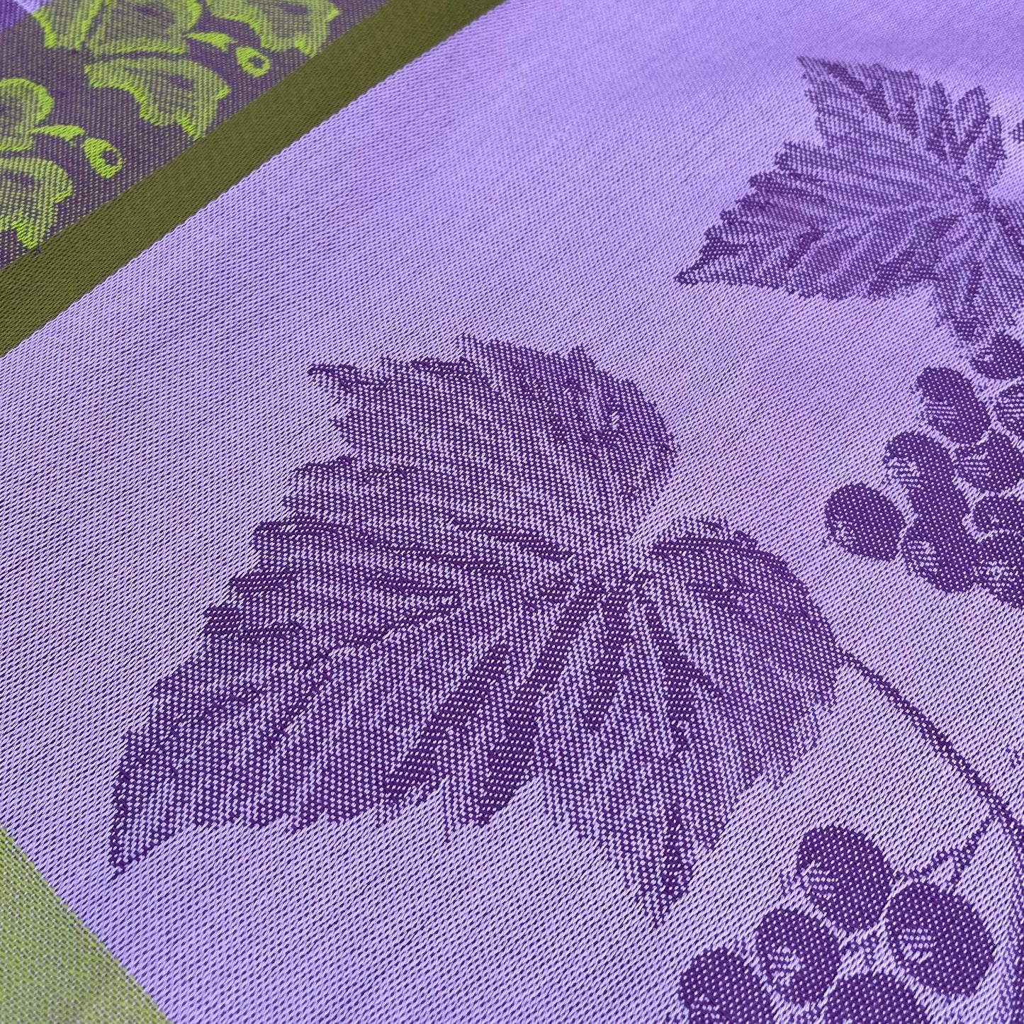 Nahaufnahme des Garnier Thiebaut Geschirrtuchs 'Les Cassis Mauve', mit einem gewebten Traubenblatt- und Traubenmuster aus mauvefarbener Baumwolle, akzentuiert mit einer grünen Bordüre und einem passenden Traubenmotiv.