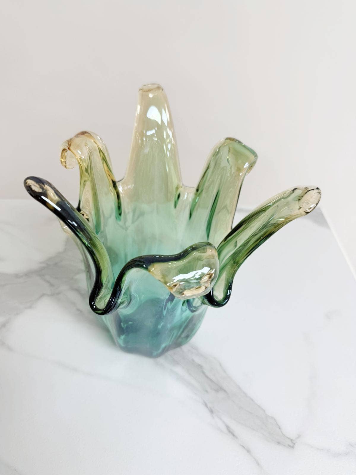 Die Murano Dream „Giusy“-Vase aus grünem Murano-Glas (H 24, Ø 18 cm) besticht durch handgefertigte, wellenförmige Kanten und setzt auf weißem Marmor vor hellem Hintergrund elegante Akzente in jedem Raum.