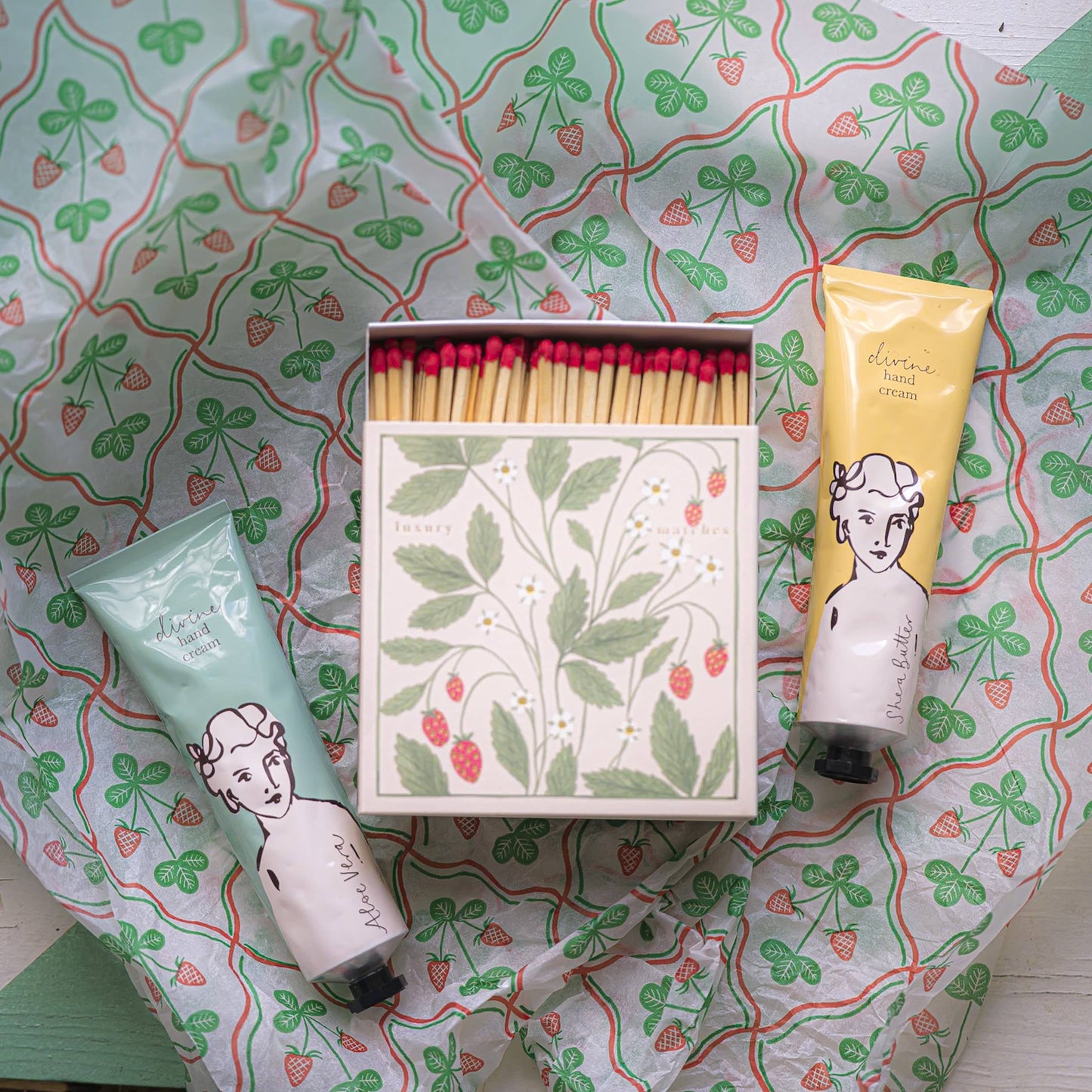 Zwei Tuben Archivist | 'Sheabutter' Handcreme von Archivist Gallery neben einer dekorativen Streichholzschachtel mit Erdbeerdesign - arrangiert auf gemustertem Papier mit grünen Blättern und roten Erdbeeren.