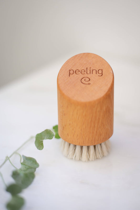 Eine Croll & Denecke Gesichtsbürste aus Holz für Peeling & Massage mit Rosshaarborsten und "Peeling"-Gravur liegt auf weißem Untergrund neben einem grünen Blatt.
