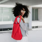 Eine Person mit lockigem Haar und Brille trägt draußen ein kariertes Oberteil, eine weiße Hose und die Notabag | „Original Red“ von Notabag über der Schulter – praktisch als Tasche und Rucksack – und blickt zurück.