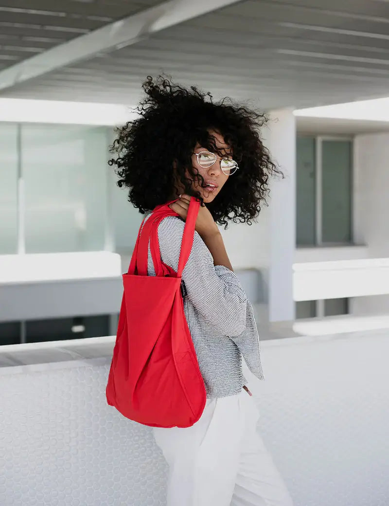 Eine Person mit lockigem Haar und Brille trägt draußen ein kariertes Oberteil, eine weiße Hose und die Notabag | „Original Red“ von Notabag über der Schulter – praktisch als Tasche und Rucksack – und blickt zurück.