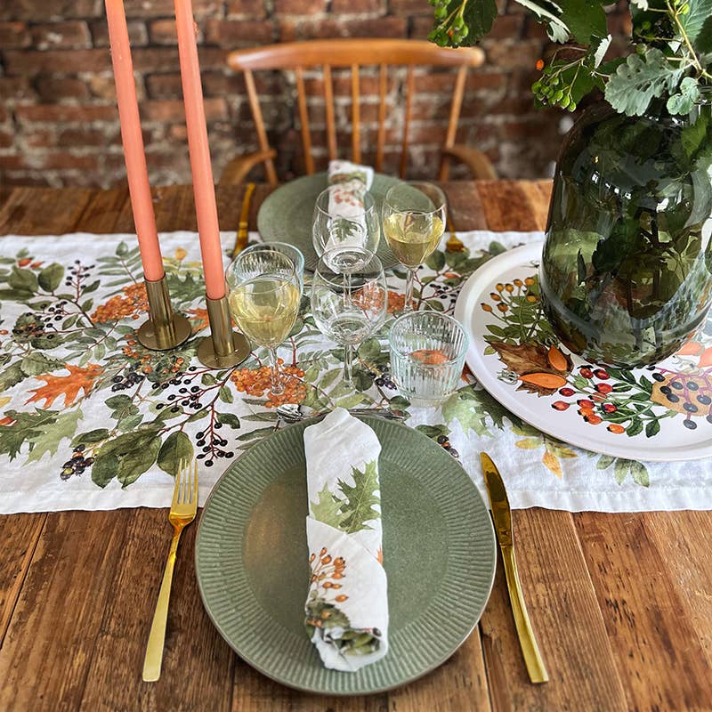 Ein für zwei Personen gedeckter Holztisch ist mit grünen Tellern, goldenem Besteck, Weingläsern, rosa Kerzen und einem blumigen Tischläufer gedeckt. Neben dem Gedeck steht eine Vase mit belaubten Zweigen auf dem Koustrupco-Serviertablett Ø 38 cm „Herbst“ und verleiht dem gemütlichen Arrangement einen eleganten Akzent.