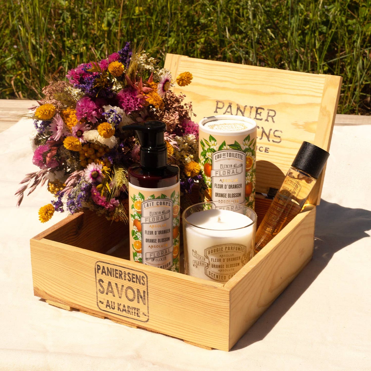 Eine hölzerne Geschenkbox mit einer nach Blumen duftenden Hautpflege, der Kerze Panier Des Sens Duftkerze | Orangenblüte 180 g und einem Wildblumenstrauß, arrangiert im Freien auf leichtem Stoff vor grünem Gras.