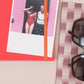 Auf dem Tinne Mia Notizbuch mit japanischem Verschluss 'Cinnamon' liegt ein Polaroidfoto von zwei Personen, eine im roten Badeanzug und eine im weißen Top. Eine Sonnenbrille und ein gestreiftes Tuch liegen in der Nähe auf der beigen Oberfläche.