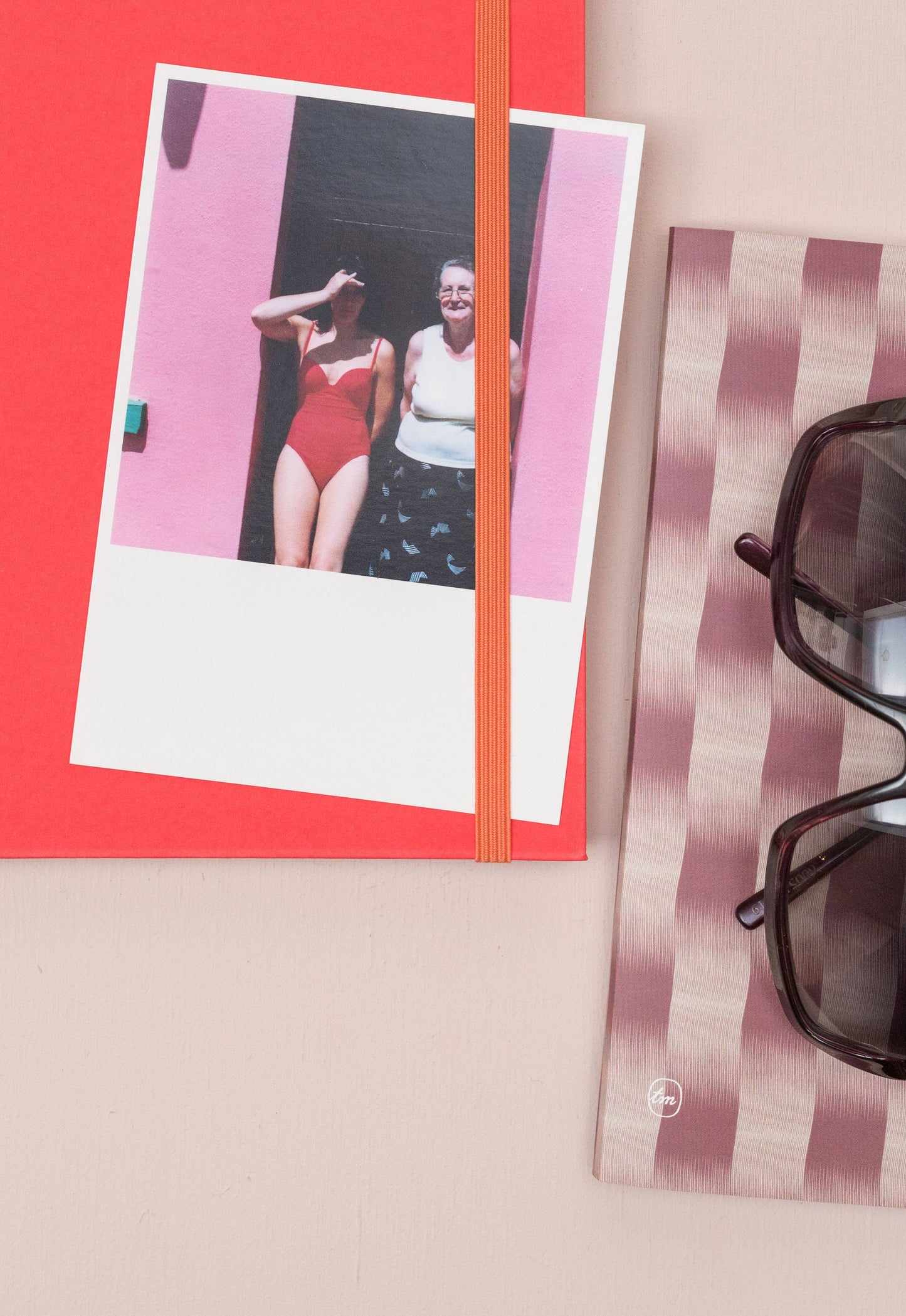 Auf dem Tinne Mia Notizbuch mit japanischem Verschluss 'Cinnamon' liegt ein Polaroidfoto von zwei Personen, eine im roten Badeanzug und eine im weißen Top. Eine Sonnenbrille und ein gestreiftes Tuch liegen in der Nähe auf der beigen Oberfläche.
