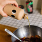Eine Hand gießt yiayia and friends Natives Olivenöl Extra mit Knoblauch 200 ml in eine Metallschale mit gehacktem Gemüse und einem Löffel auf einem karierten Tisch, mit Flaschen im Hintergrund - ideal für die mediterrane Küche.