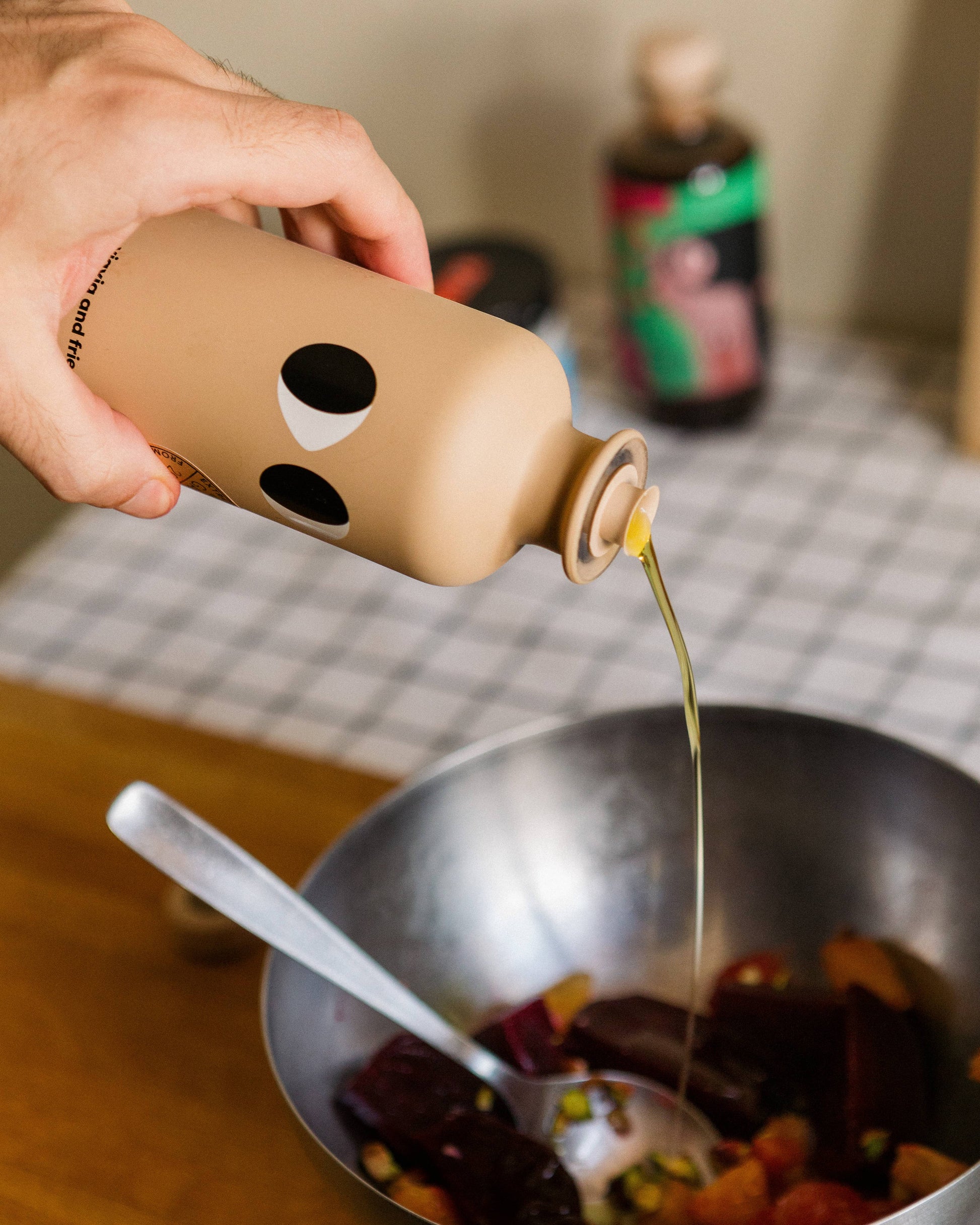 Eine Hand gießt yiayia and friends Natives Olivenöl Extra mit Knoblauch 200 ml in eine Metallschale mit gehacktem Gemüse und einem Löffel auf einem karierten Tisch, mit Flaschen im Hintergrund - ideal für die mediterrane Küche.