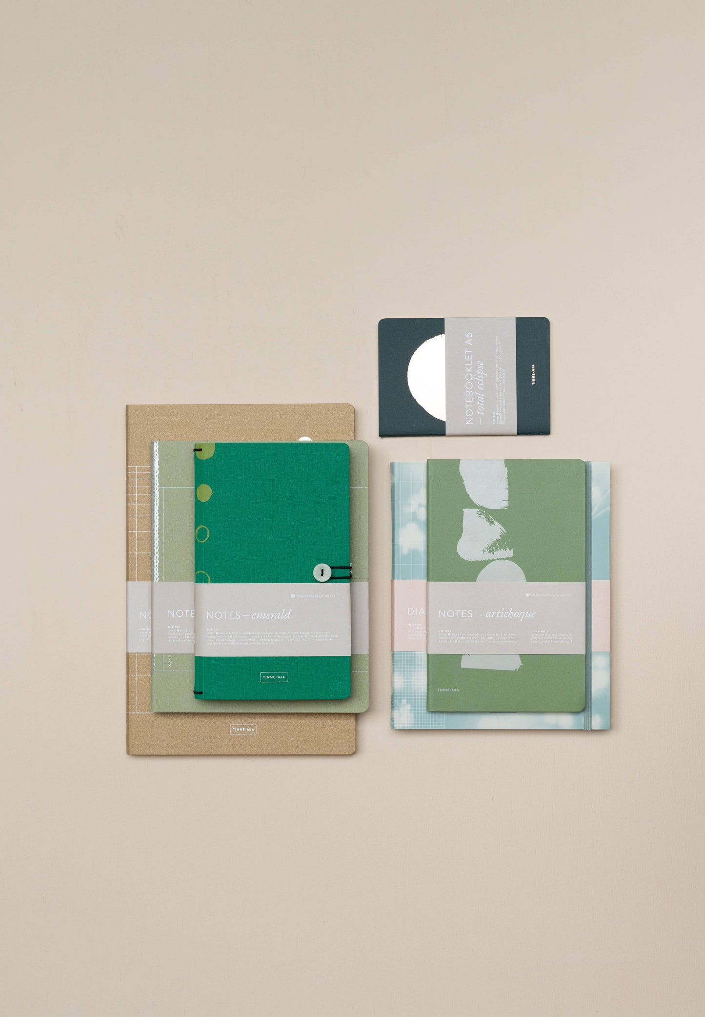 Ein übersichtliches Tinne Mia Notizbuch mit Knopf 'Leinen Smaragd' von Tinne Mia, FSC-zertifiziert, liegt auf einer beigen Unterlage - ideal für die Verwendung als Tagebuch oder Bullet Journal. Umgeben ist es von weiteren Notizbüchern und einer kleinen grauen Karte mit weißem Kreismuster.