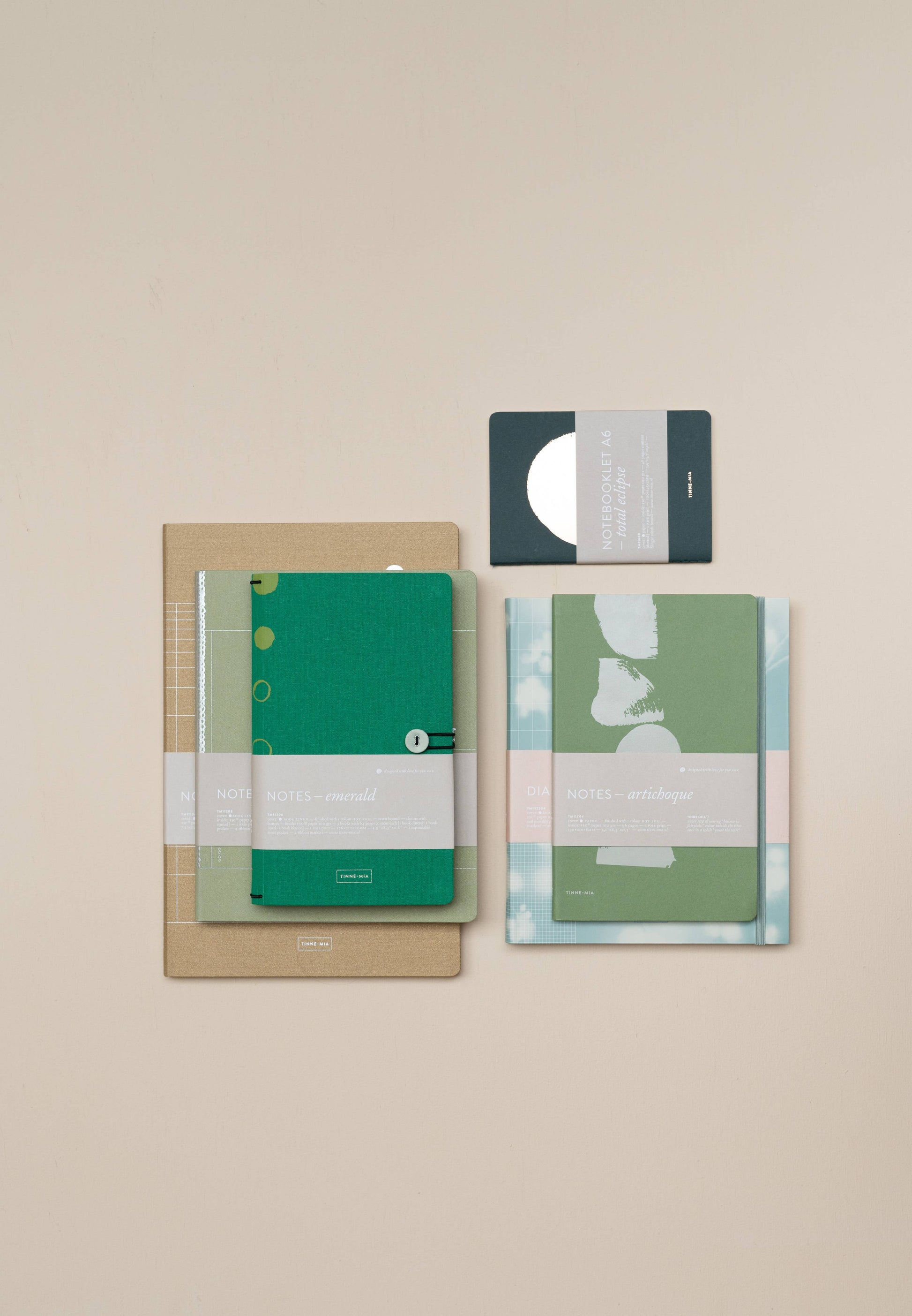 Ein übersichtliches Tinne Mia Notizbuch mit Knopf 'Leinen Smaragd' von Tinne Mia, FSC-zertifiziert, liegt auf einer beigen Unterlage - ideal für die Verwendung als Tagebuch oder Bullet Journal. Umgeben ist es von weiteren Notizbüchern und einer kleinen grauen Karte mit weißem Kreismuster.
