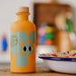 Eine gelbe 200-ml-Flasche Yiayia and friends | Weißer Balsamico-Essig mit großen blaugrünen Kreisen und schwarzen Punkten auf dem Etikett steht auf einer hölzernen Unterlage neben einem Teller mit Speisen vor einem unscharfen Küchenhintergrund, ideal für eine Vinaigrette.