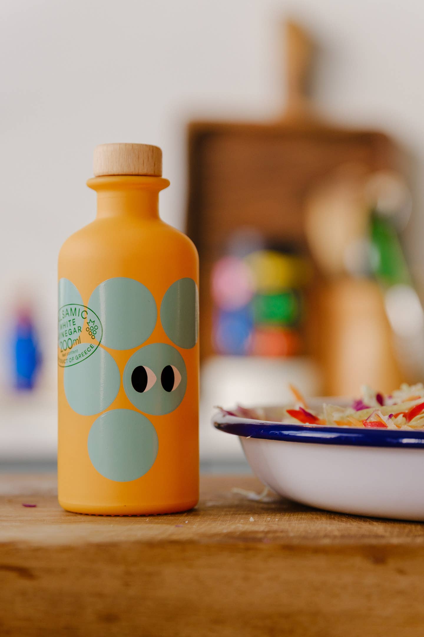 Eine gelbe 200-ml-Flasche Yiayia and friends | Weißer Balsamico-Essig mit großen blaugrünen Kreisen und schwarzen Punkten auf dem Etikett steht auf einer hölzernen Unterlage neben einem Teller mit Speisen vor einem unscharfen Küchenhintergrund, ideal für eine Vinaigrette.