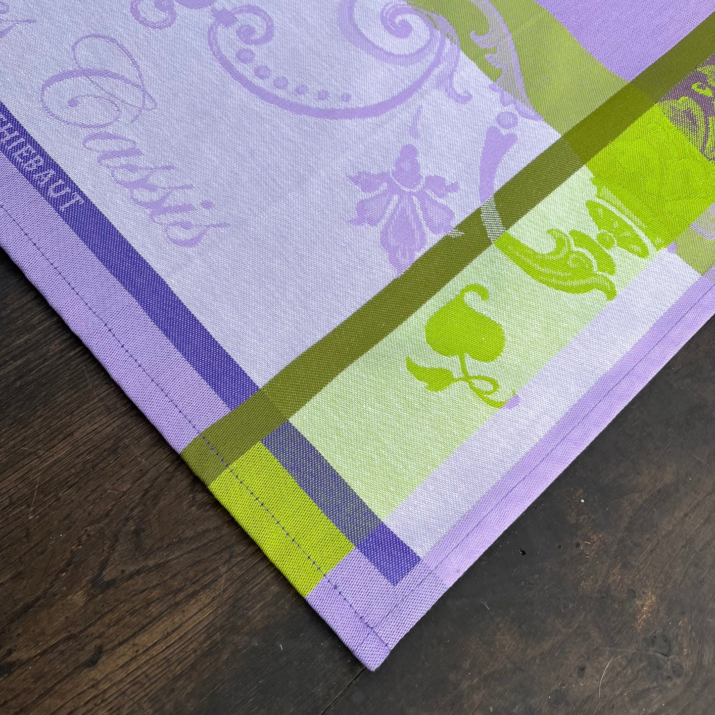 Eine Nahaufnahme des Garnier Thiebaut Geschirrtuchs "Les Cassis Mauve" mit hellvioletten, grünen und limonenfarbenen Streifen mit floralen Mustern und seinem aufgedruckten Namen, ausgestellt auf einer dunklen Holzoberfläche.