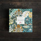 Eine quadratische Koustrupco-Schachtel mit einem Blumen- und Vogelmotiv von William Morris in Grün-, Gold-, Creme- und Oxford-Rot-Tönen ruht auf einer hölzernen Unterlage und trägt die Aufschrift "Koustrupco | Seidenschal Morris Oxford Rot 50 x 50 cm".