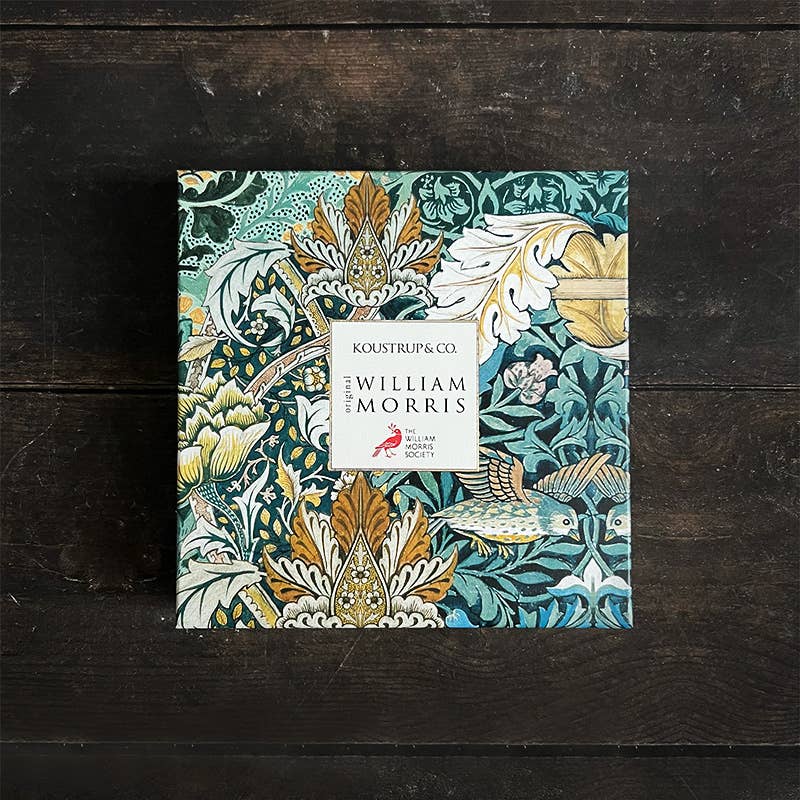 Eine quadratische Koustrupco-Schachtel mit einem Blumen- und Vogelmotiv von William Morris in Grün-, Gold-, Creme- und Oxford-Rot-Tönen ruht auf einer hölzernen Unterlage und trägt die Aufschrift "Koustrupco | Seidenschal Morris Oxford Rot 50 x 50 cm".
