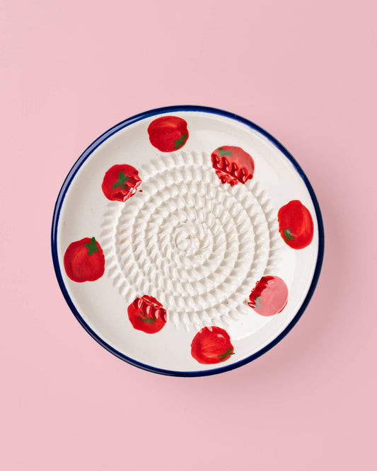 Die Alfar Tierra Cocida | Kleine Keramik-Knoblauchreibe „Pomodoro“ von Alfar Tierra Cocida, eine handgefertigte weiße Knoblauchreibe aus Spanien mit spiralförmiger Reibfläche und roten Tomatenmotiven am Rand, liegt auf einem pinkfarbenen Hintergrund.