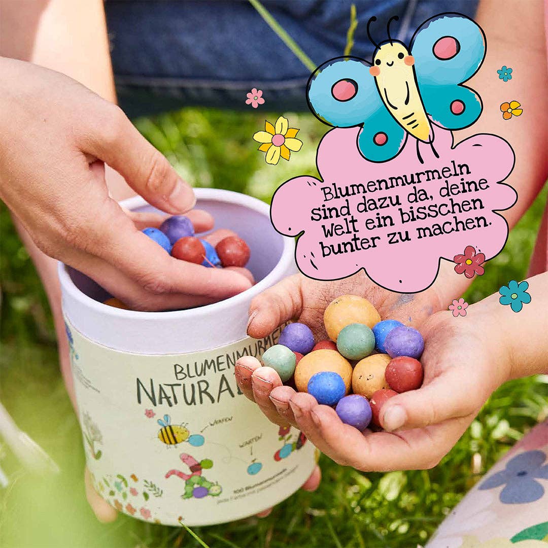 Hände, die bunte Blumenmurmeln von Stadtgärtner Saatgut halten | Blumenmurmeln „Naturabenteuer“ 100er-Dose, daneben ein passender Behälter. Ein illustrierter Schmetterling und eine verspielte Sprechblase verkünden: „Blumenmurmeln sind dazu da, deine Welt ein bisschen bunter zu machen.“ Fröhliche Blumen umrahmen die Szene und laden die Kleinen zum Entdecken ein – das perfekte Naturabenteuer und ein nachhaltiges Geschenk von Stadtgärtner für Kinder.