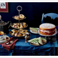 Das Tischset "London Lovestory" vom Hölker Verlag mit blauem Tuch, Stufentablett mit Scones, Biskuit, Gurkensandwiches, Teekanne, Teetasse, Blumen in einer Vase und blauem Stuhl bringt Londoner Romantik in die Teestunde.