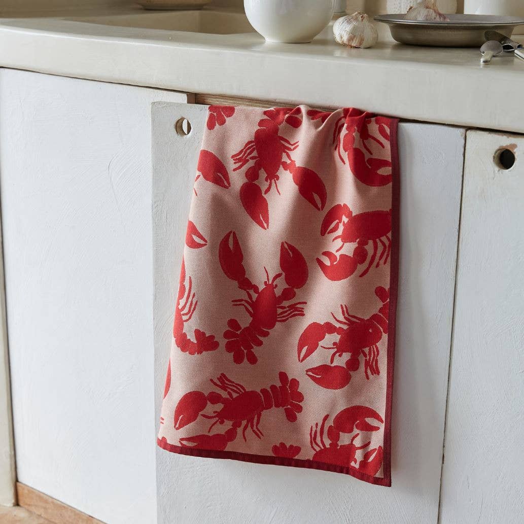 Ein rotes Geschirrtuch „Bonne pêche“ von Sylvie Thiriez mit roten Hummermotiven hängt über einer weißen Schranktür neben einer Küchenarbeitsplatte, auf der eine Knoblauchknolle und eine weiße Schüssel stehen.