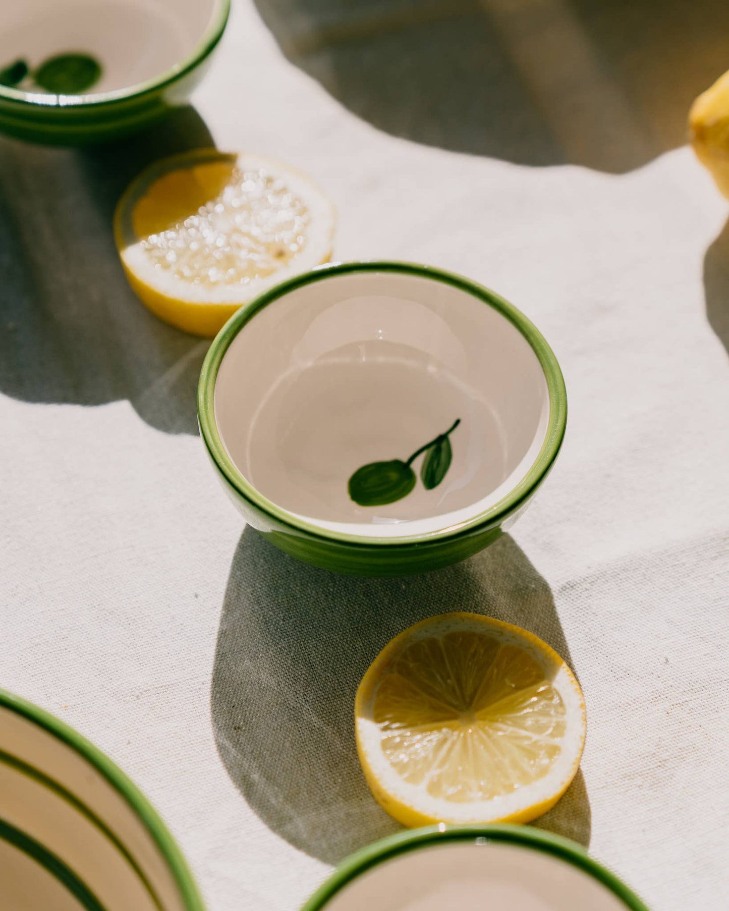 Eine kleine Keramikschale „Olive“ von Alfar Tierra Cocida | Handgefertigte kleine Keramikschale von Alfar Tierra Cocida mit grünem Rand und Blattmotiv steht auf einer Tischdecke neben Zitronenscheiben. Handgefertigt in Spanien, fängt die mediterrane Keramikschale das Sonnenlicht ein und wirft sanfte Schatten auf die Szenerie.