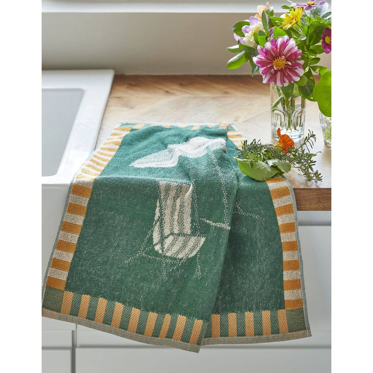 Das Sylvie Thiriez Frottee-Handtuch 'Sonnenliegen' 50x50 cm mit grünen und goldenen Streifen und weißem Vogelmotiv ruht auf einer hölzernen Küchenzeile neben einer Glasvase mit bunten Blumen. OEKO-TEX-zertifiziert für ein sicheres Gefühl.