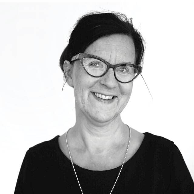 Eine Frau mit zurückgebundenen dunklen Haaren, Brille und Halskette lächelt in einem dunklen Oberteil vor weißem Hintergrund in die Kamera. Das Schwarz-Weiß-Foto fängt die Schlichtheit des biologisch abbaubaren Schwammtuchs "Kranz aus Stiefmütterchen" von More Joy Home ein.