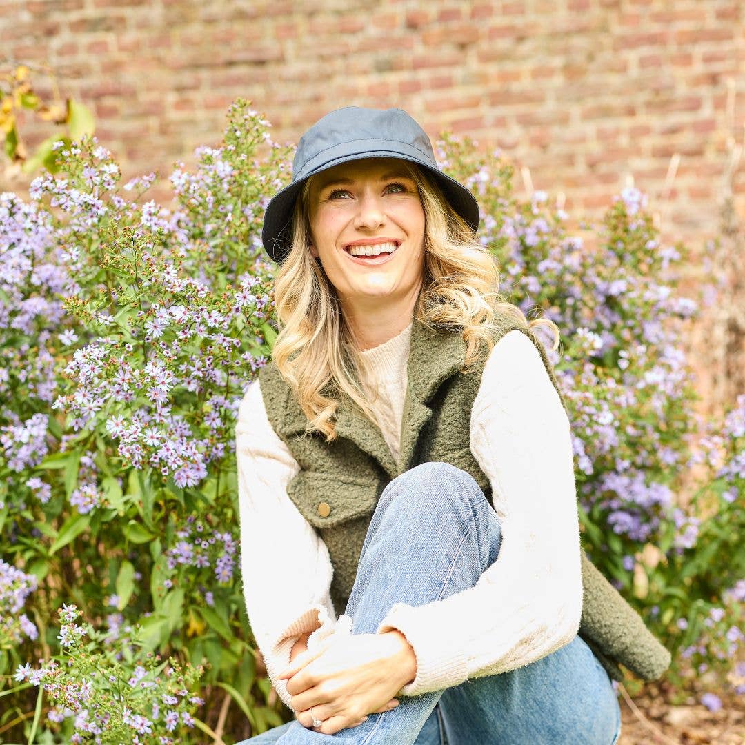 Eine Frau trägt einen Hut mit gewachstem Krempen von Tweedmill Textiles, dazu eine grüne Weste und Jeans. Sie sitzt lächelnd vor lila Blumen, im Hintergrund ist eine Backsteinmauer zu sehen.
