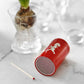 Ein kleiner roter tragbarer Lautsprecher mit einem weißen Pferdelogo liegt auf der Seite auf einer Marmorfläche neben der Solstickan Design | Streichholzdose Rot und einer Glasvase mit einer sprießenden Glühbirne.