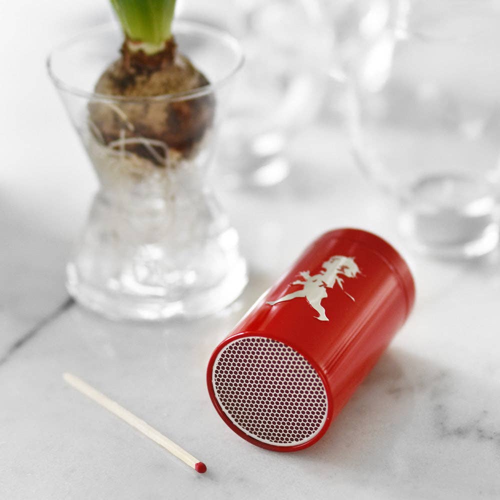 Ein kleiner roter tragbarer Lautsprecher mit einem weißen Pferdelogo liegt auf der Seite auf einer Marmorfläche neben der Solstickan Design | Streichholzdose Rot und einer Glasvase mit einer sprießenden Glühbirne.