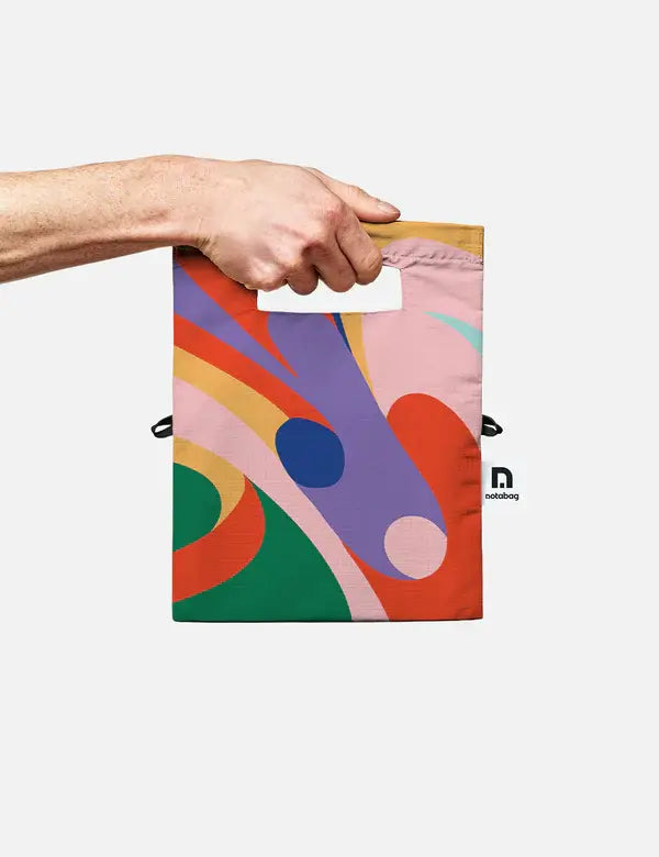 Eine Hand hält die Notabag | 'Flow' Leichte Umhängetasche von Notabag, eine farbenfrohe rechteckige Tote mit ausgeschnittenem Henkel, kühnen abstrakten Mustern, Seitenlabel und verstellbarem Gurt für vielseitige Trageoptionen.