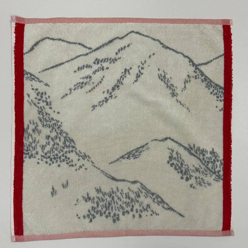 Das Sylvie Thiriez Küchenhandtuch 'Grandeur nature' zeigt ein weißes Baumwolldesign mit grauen, skizzenhaften Berg- und Baummotiven, die mit roten und rosafarbenen Bordüren abschließen.