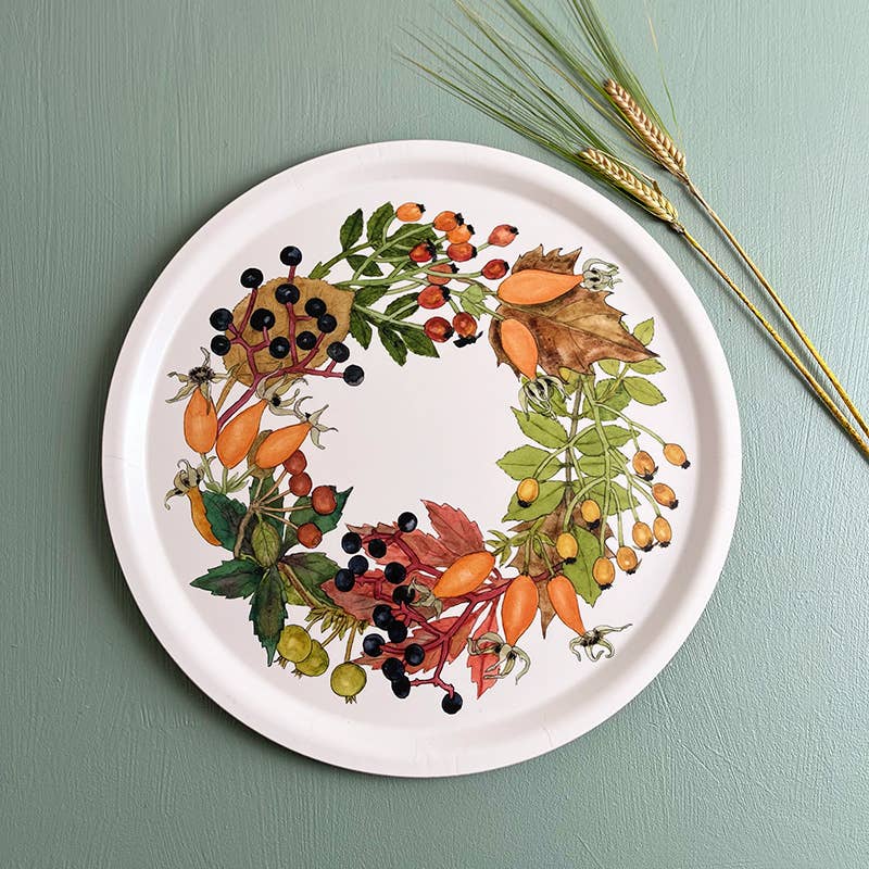 Ein rundes Serviertablett von Koustrupco, das „Serviertablett Ø 38 cm Herbst“, besticht durch ein farbenfrohes botanisches Kranzmotiv mit Beeren, Hagebutten und Blättern. Wie bei einem dekorativen Tablett liegen drei Weizenhalme neben dem Koustrupco-Tablett auf einer grünen Unterlage.