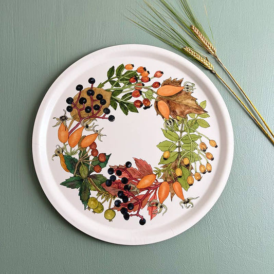 Ein rundes Serviertablett von Koustrupco, das „Serviertablett Ø 38 cm Herbst“, besticht durch ein farbenfrohes botanisches Kranzmotiv mit Beeren, Hagebutten und Blättern. Wie bei einem dekorativen Tablett liegen drei Weizenhalme neben dem Koustrupco-Tablett auf einer grünen Unterlage.