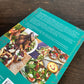 Das Bild zeigt die Rückseite von Das Kochbuch | Ottolenghi von DK auf einem Holztisch, mit deutschem Text und Fotos von Gerichten wie Lammkoteletts, Salaten und Obst-Sahne-Desserts - eine klassische Ottolenghi-Inspiration.
