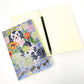 Ein offenes leeres Notizbuch mit einem schwarzen Stift liegt neben dem geschlossenen Art Angels Notizbuch 'Wally Dogs and Tulpen' von Angie Lewin, das ihr farbenfrohes Blumen- und Wally Dogs-Design zeigt, alles auf einer weißen Oberfläche.