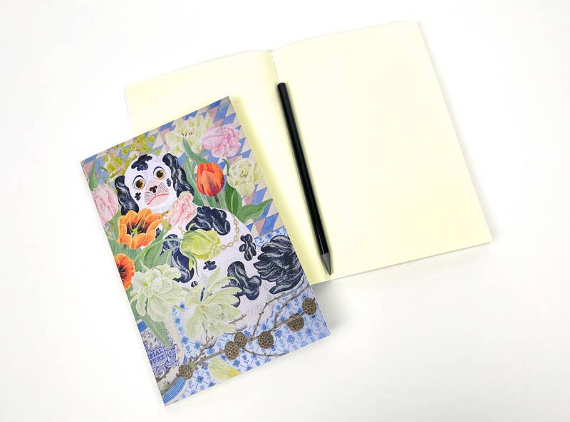 Ein offenes leeres Notizbuch mit einem schwarzen Stift liegt neben dem geschlossenen Art Angels Notizbuch 'Wally Dogs and Tulpen' von Angie Lewin, das ihr farbenfrohes Blumen- und Wally Dogs-Design zeigt, alles auf einer weißen Oberfläche.
