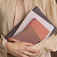 Eine Person in cremefarbener Bluse hält mit beiden Armen mehrere zartfarbige Notizbücher und Mappen, darunter das Tinne Mia Notizbuch mit Knopf 'Leinen Rose Dawn', ein rosa Bullet Journal und lilafarbene Gegenstände dicht vor der Brust.