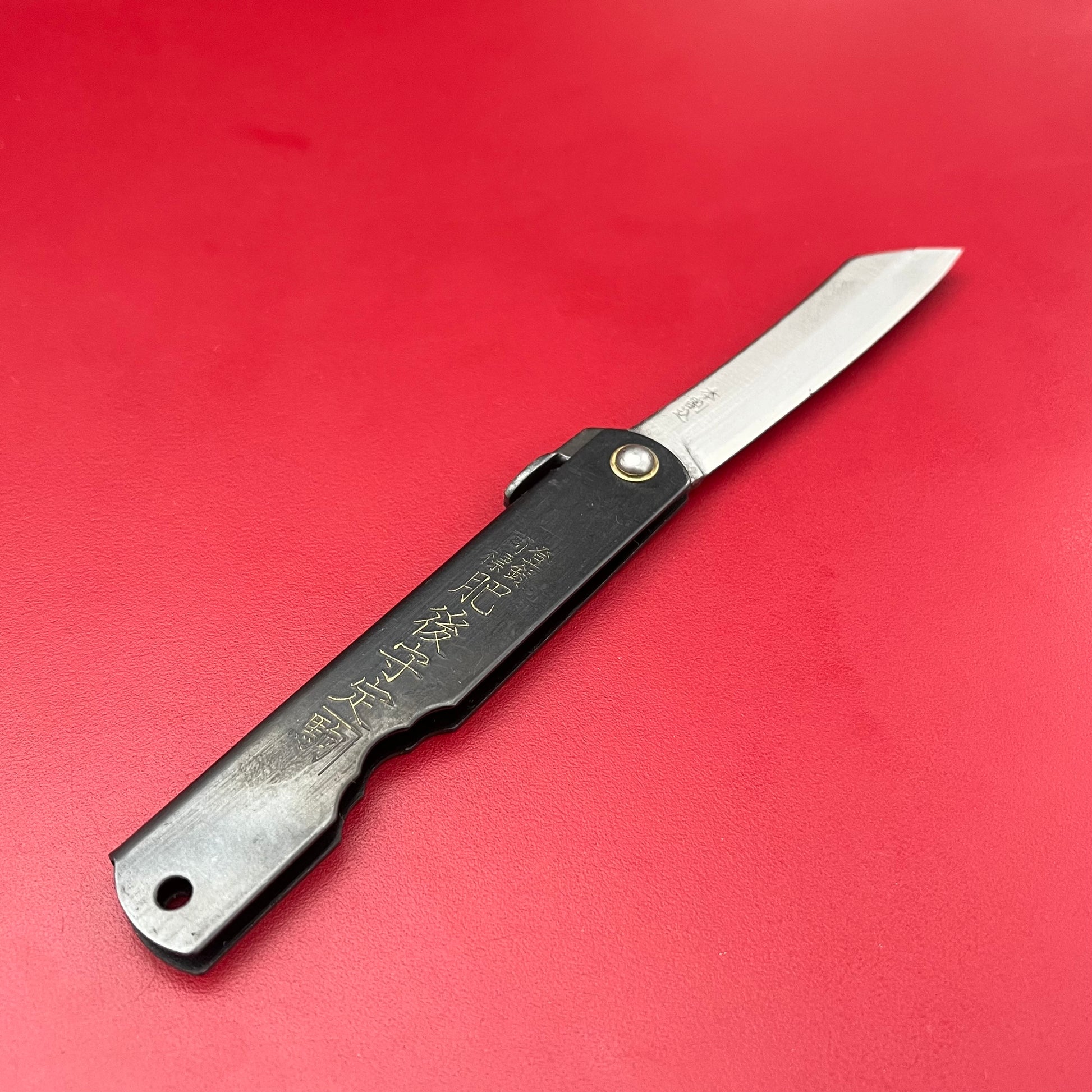 Ein Niwaki 'SK Higonokami Folding Knife' Klappmesser mit schwarzem Griff und eingraviertem Goldtext liegt geöffnet auf einer leuchtend roten Fläche, deren Klinge das Licht teilweise reflektiert.