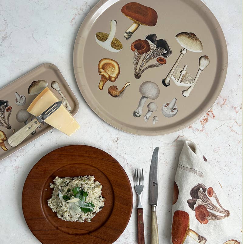 Auf einer mit Pilzmotiven verzierten Serviette liegt ein Holzteller mit Pilzrisotto, einer Gabel und einem Messer. Daneben liegen Käse und ein Messer neben zwei Tabletts „Pilz“ von Koustrupco | Serviertablett 32 x 15 cm, die jeweils mit verschiedenen Pilzen auf weißem Hintergrund verziert sind.