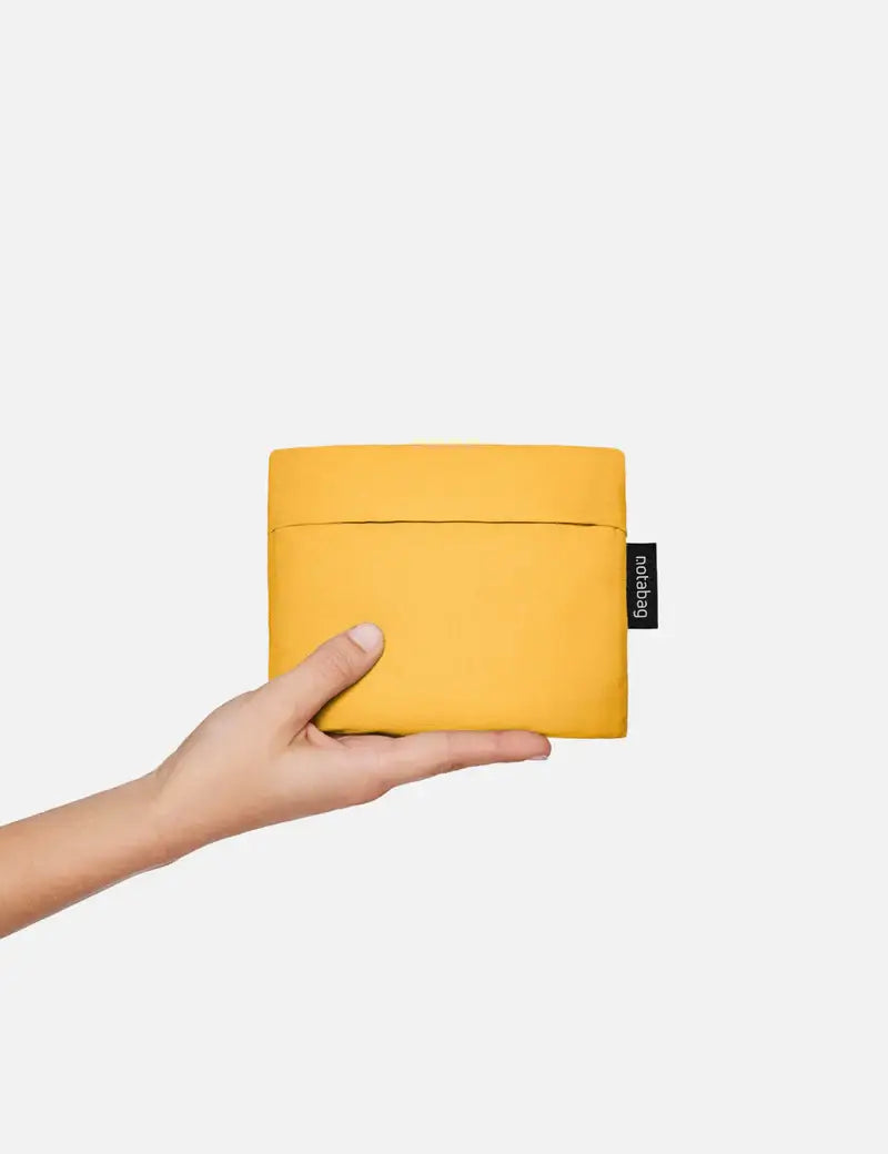 Eine Hand hält eine kleine, rechteckige, leuchtend gelbe Notabag „Original Golden“ – eine Kombination aus Tragetasche und Rucksack – mit einem schwarzen Notabag-Etikett vor einem schlichten weißen Hintergrund.