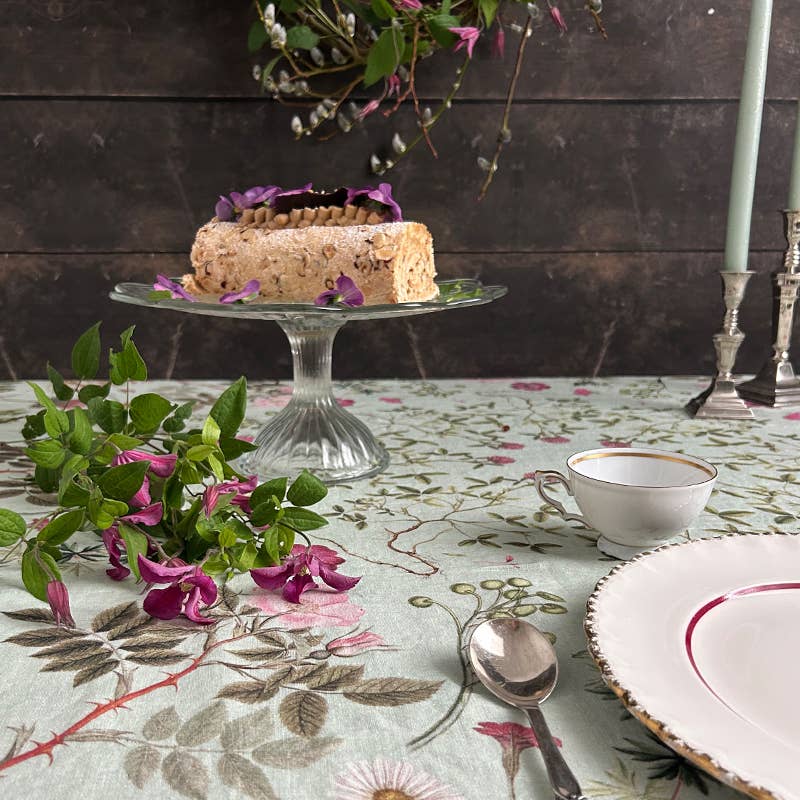 Ein kleiner Kuchen mit violetten Blumen steht auf einem Glasständer auf der Tischdecke „Flora Danica Atlas“ von Koustrupco (145 x 220 cm), umgeben von botanischer Tischdekoration, darunter eine Tasse, eine Untertasse, ein Löffel sowie hohe Kerzen mit Blüten und Grün.