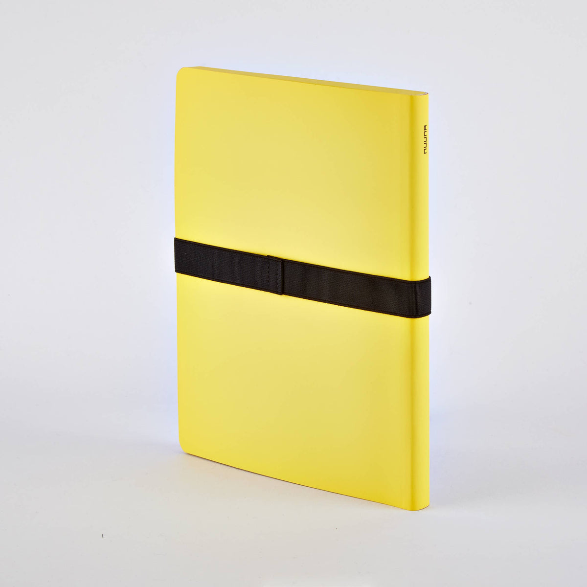 Ein geschlossenes gelbes Nuuna-Notizbuch „Not White Yellow“ L Light A5+ aus dem Notizbuch-Set mit Gelstift steht aufrecht vor einem weißen Hintergrund und wird in der Mitte von einem horizontalen schwarzen Gummiband zusammengehalten.