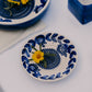 Die Alfar Tierra Cocida | Kleine Keramik-Knoblauchreibeplatte „Nerja“, blau und traditionell bemalt, eine kleine Keramik-Knoblauchreibeplatte von Alfar Tierra Cocida mit blauen Blattmustern, auf der eine gelbe Margerite mit grünem Stiel zu sehen ist. Sie steht auf einem weißen Untergrund, auf dem auch ein Teil einer weiteren Schale und eine Vase zu sehen sind.