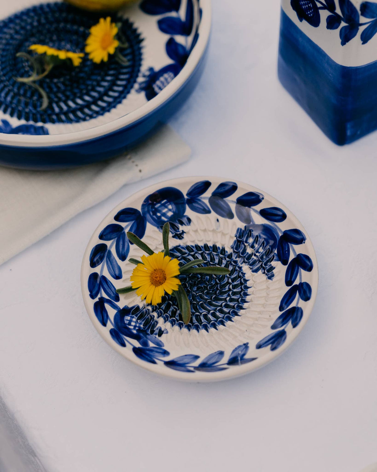 Die Alfar Tierra Cocida | Kleine Keramik-Knoblauchreibeplatte „Nerja“, blau und traditionell bemalt, eine kleine Keramik-Knoblauchreibeplatte von Alfar Tierra Cocida mit blauen Blattmustern, auf der eine gelbe Margerite mit grünem Stiel zu sehen ist. Sie steht auf einem weißen Untergrund, auf dem auch ein Teil einer weiteren Schale und eine Vase zu sehen sind.