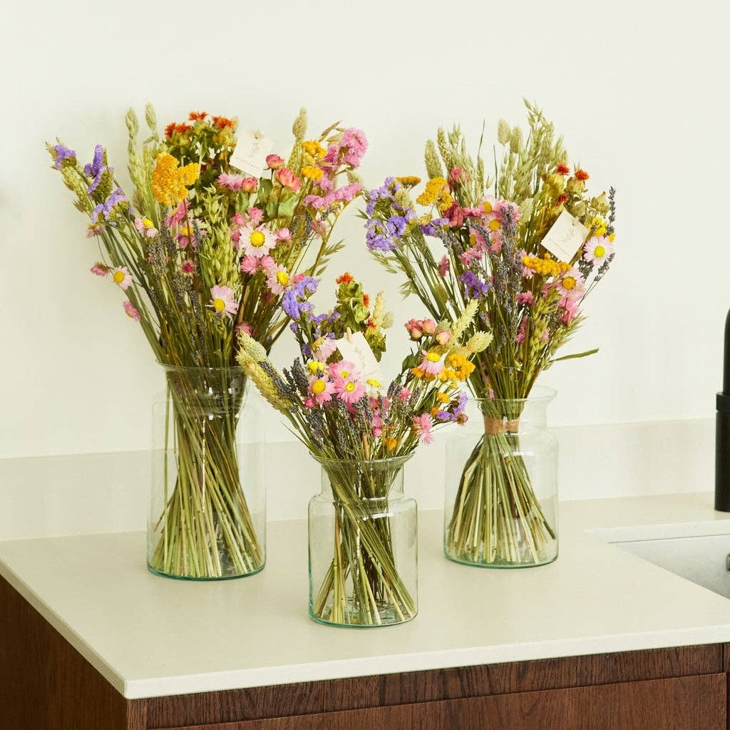 Vier Glasvasen, gefüllt mit bunten Trockenblumen, darunter der Strauß „Feldblumenstrauß“ in den Größen S und M aus der Kollektion „Wildflowers“ von Floriette, stehen auf einer weißen Arbeitsplatte neben einem dunklen Holzschrank und einer schwarzen Küchenarmatur.